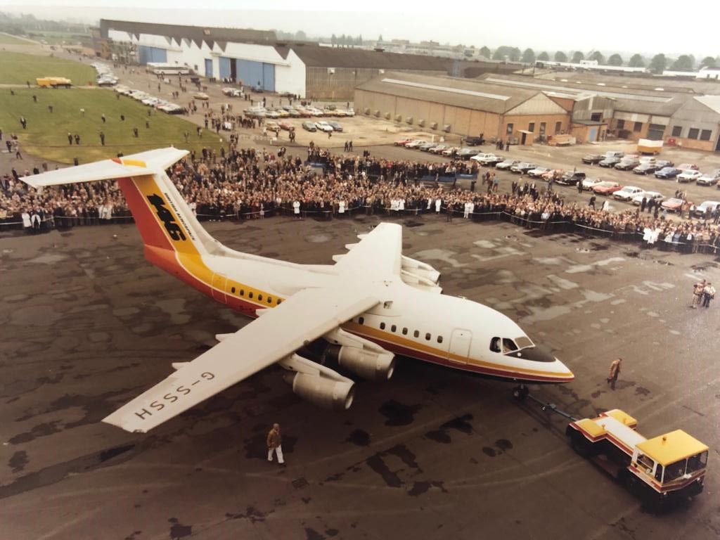 Old photo of the first BAe 146-100 aircraft, registration G-SSSH. It's painted white with a yellow and a red stripe down the fuselage and is shown on a tarmac area being displayed to a crowd of people.